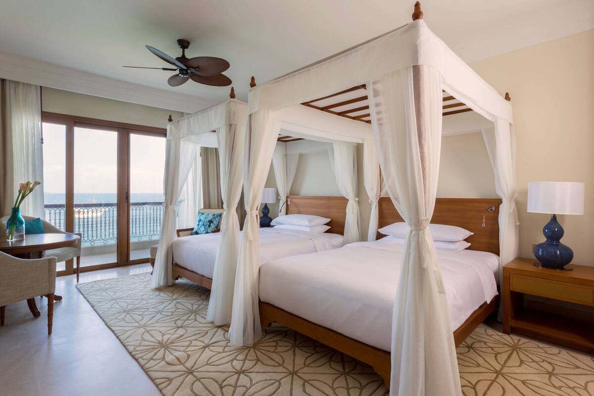 A hotel room with a four-poster bed, a desk with a chair, a nightstand, and a view of the sea through large windows.