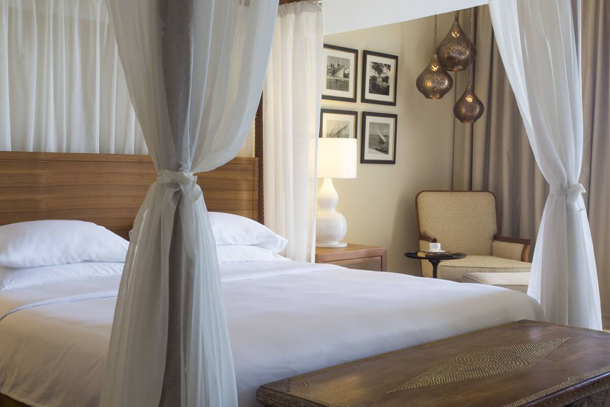 A hotel room with a large bed, a wooden headboard, a bedside table with a lamp, a chair, and a small table with a cup.