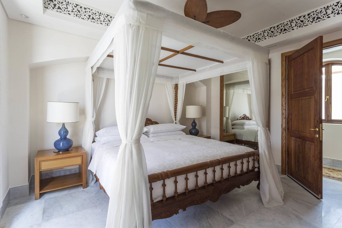 A four-poster bed with white linens, a wooden nightstand with a blue lamp, and a wooden door.