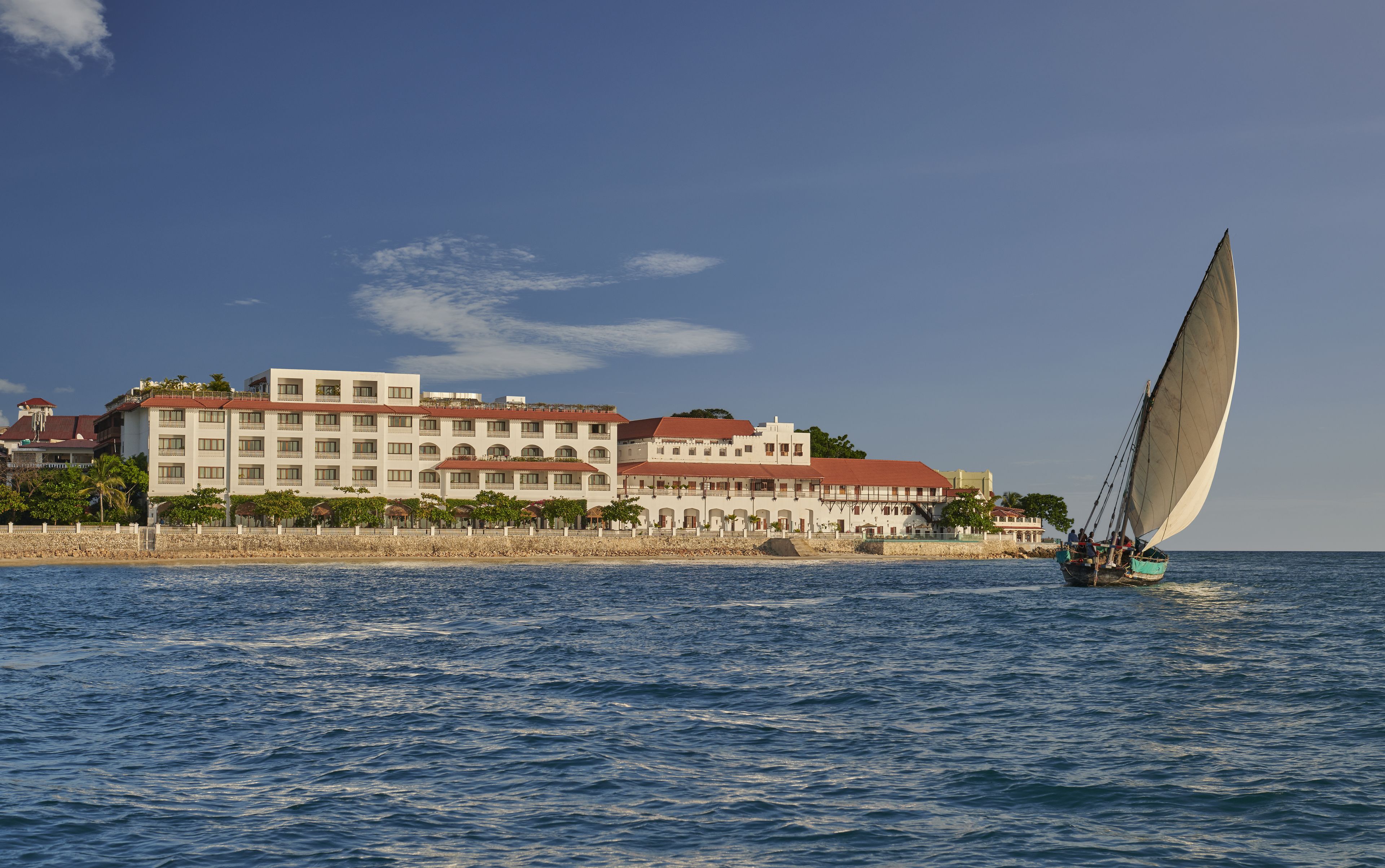 A coastal hotel with multiple stories, a beach in the foreground, and a sailboat on the water.