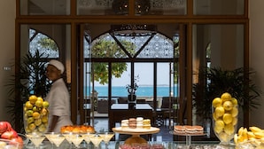 Een buffet met diverse desserts en fruit, een persoon op de achtergrond, en uitzicht op de zee door grote ramen.