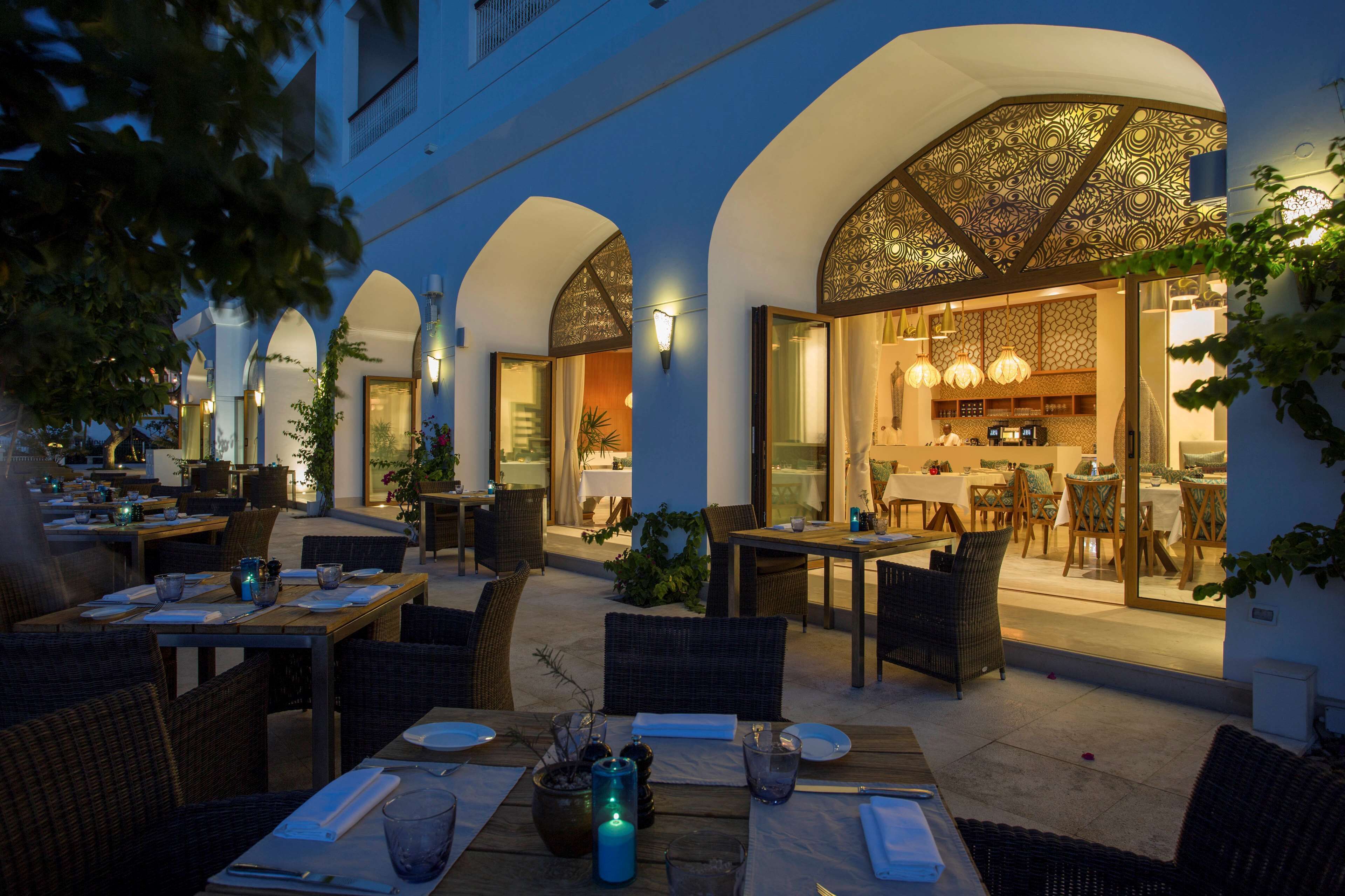 An outdoor dining area with tables and chairs, a kitchen visible through an open doorway, and arched architectural features.