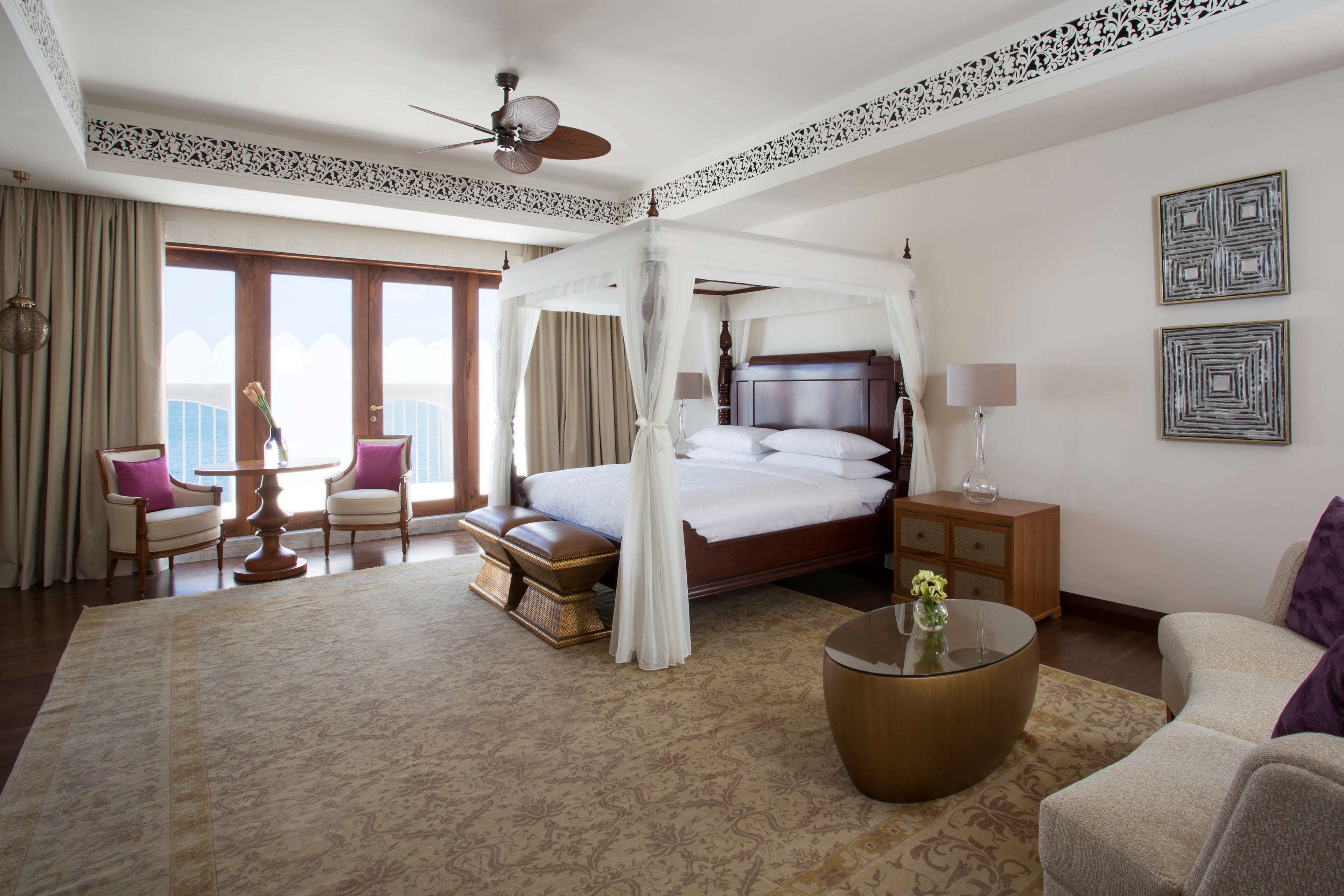 A spacious hotel room with a large bed, a sitting area with a table and chairs, a balcony with curtains, and a ceiling fan.