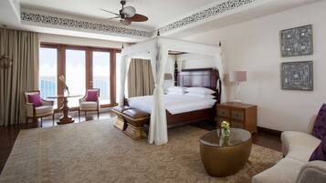 A spacious hotel room with a large bed, a sitting area with a table and chairs, a balcony with curtains, and a ceiling fan.