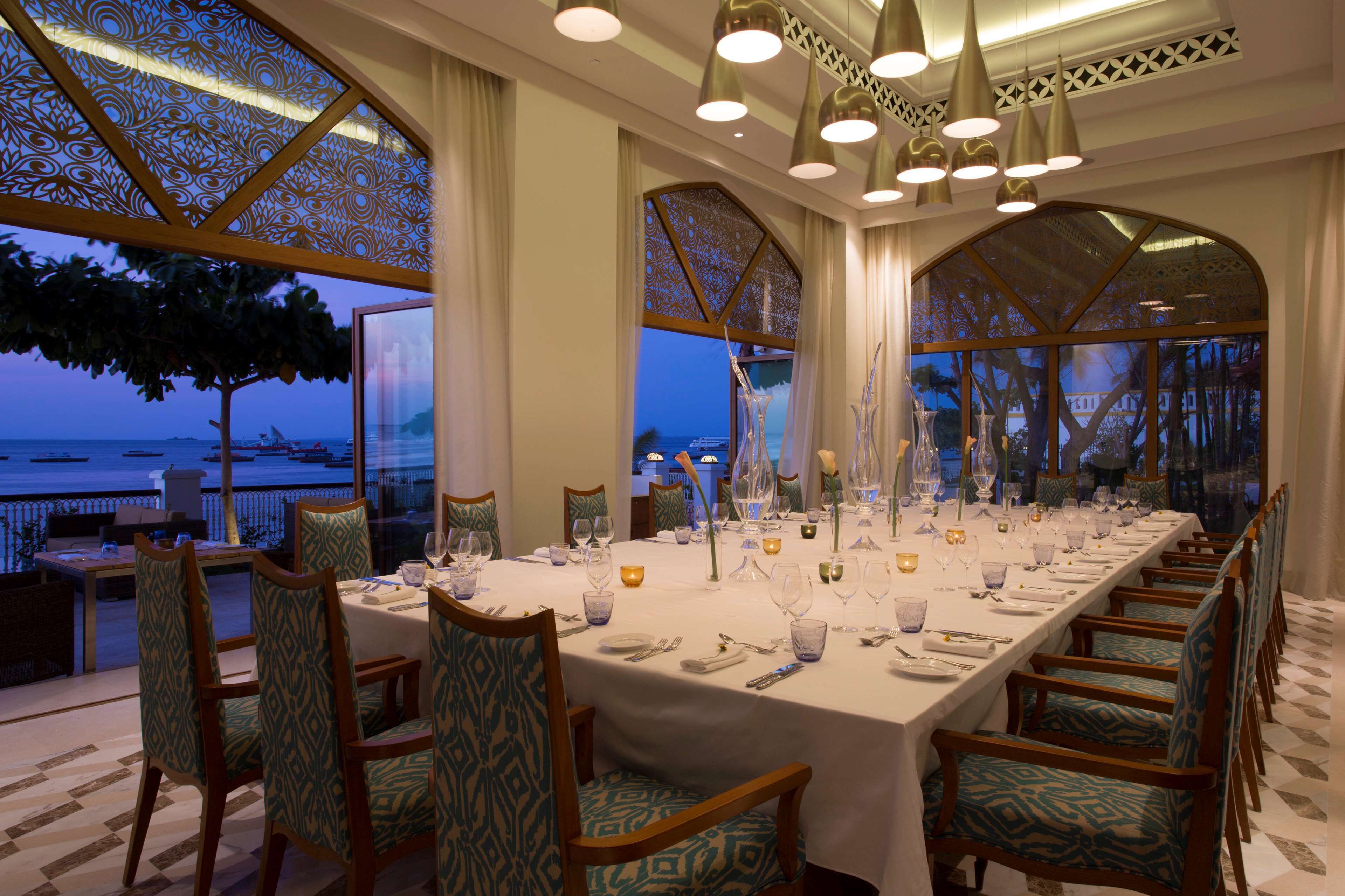 A long dining table set for a meal with chairs, a view of the sea, and decorative lighting.