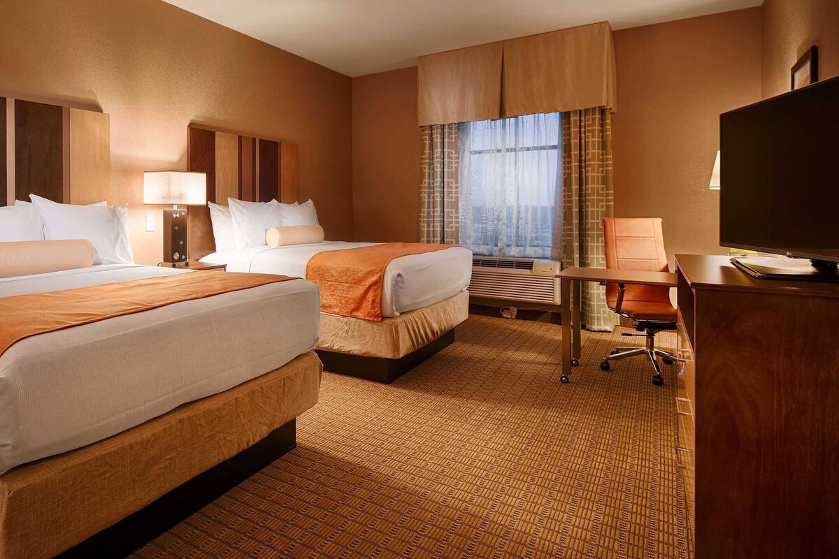 A hotel room with two beds, a desk, a chair, a TV, and a window with curtains.