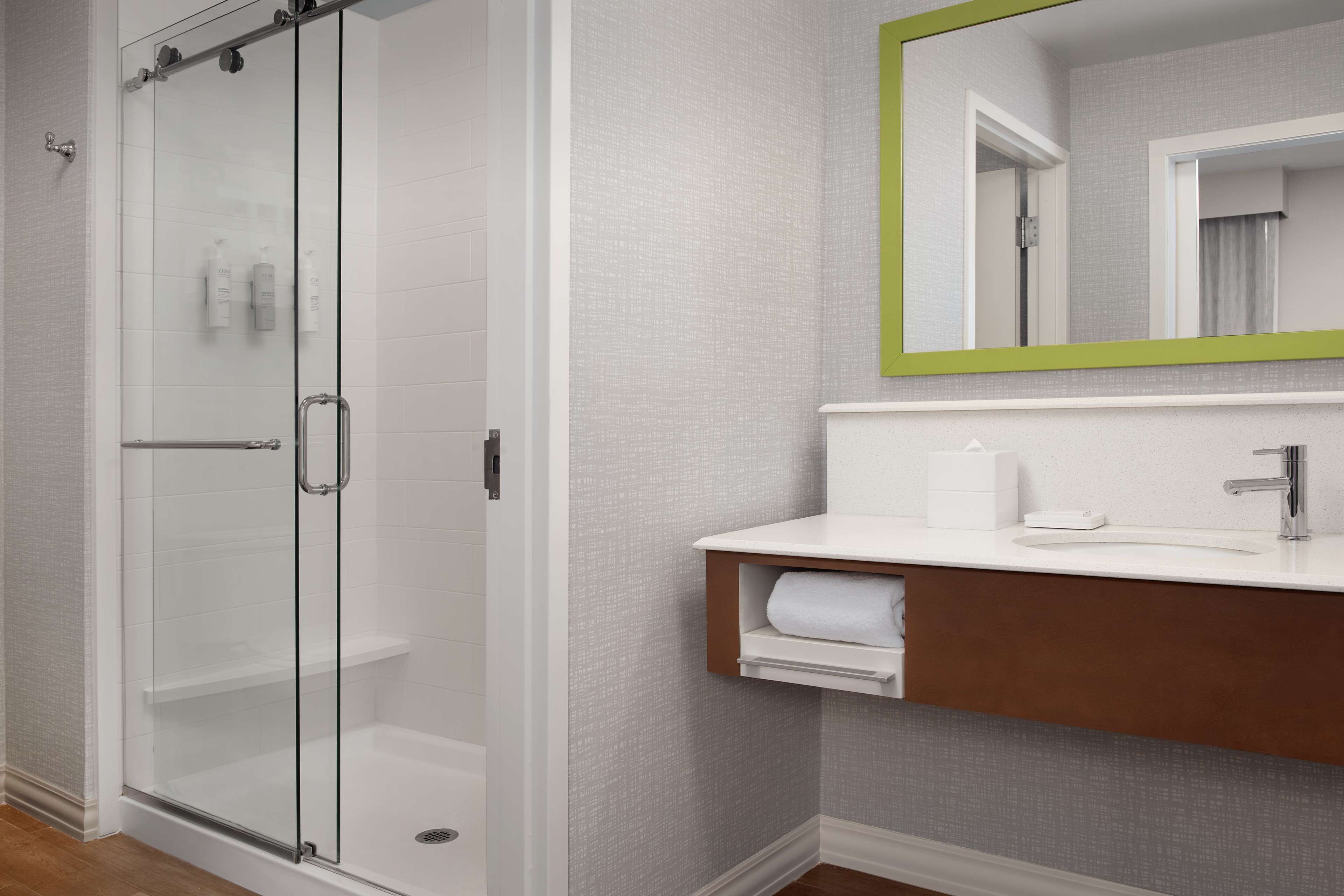 A bathroom with a glass shower enclosure, a white sink with a wooden vanity, and a mirror with a green frame.