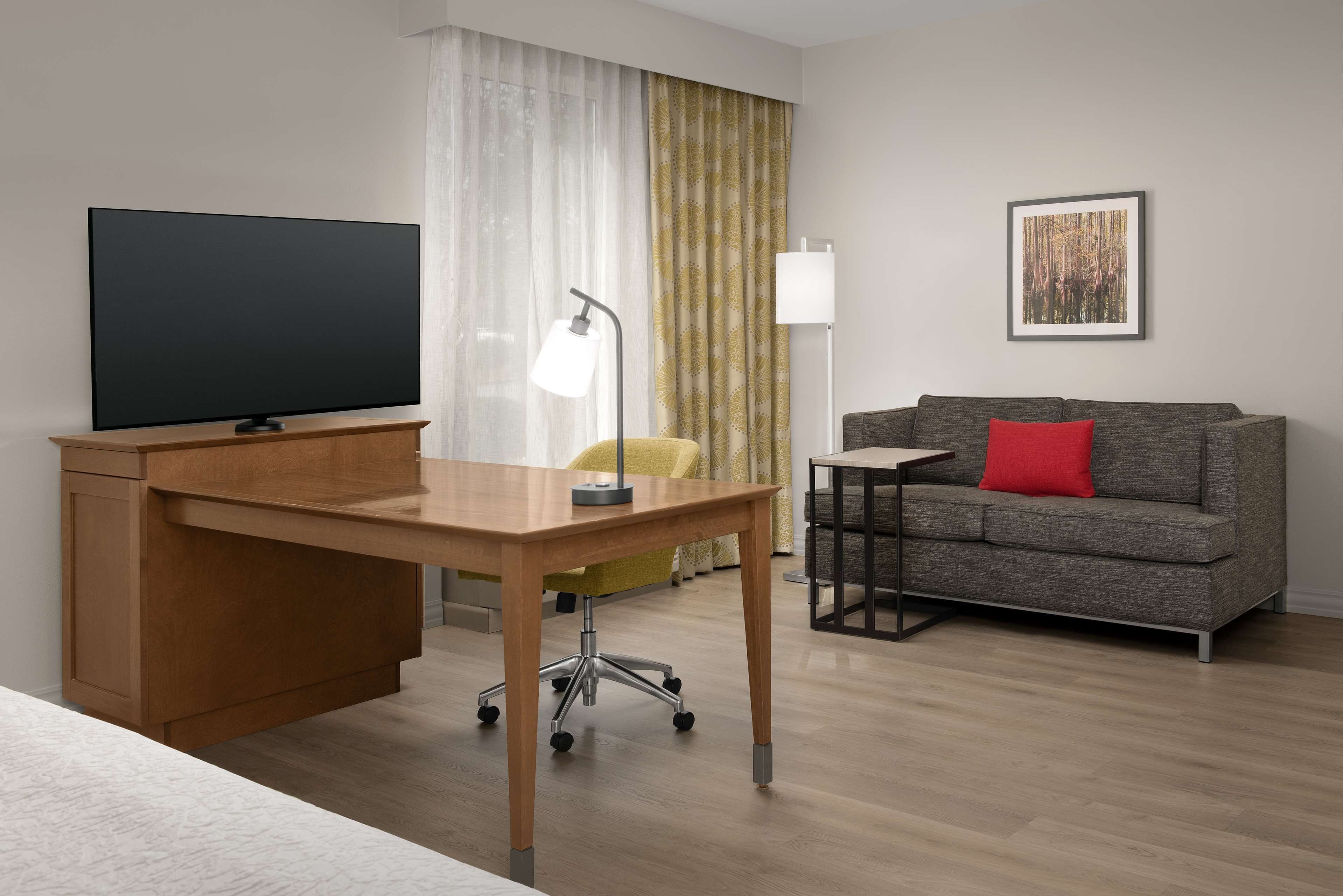 Una habitación de hotel moderna con un escritorio de madera, un televisor de pantalla plana, una silla amarilla, un sofá gris con una almohada roja y un cuadro en la pared.
