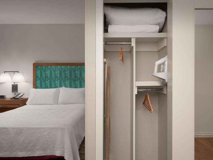 Ein Hotelzimmer mit einem ordentlich bezogenen Bett, einem Nachttisch mit Lampe, einem Kleiderschrank mit Kleidung und einem Spiegel an der Wand.