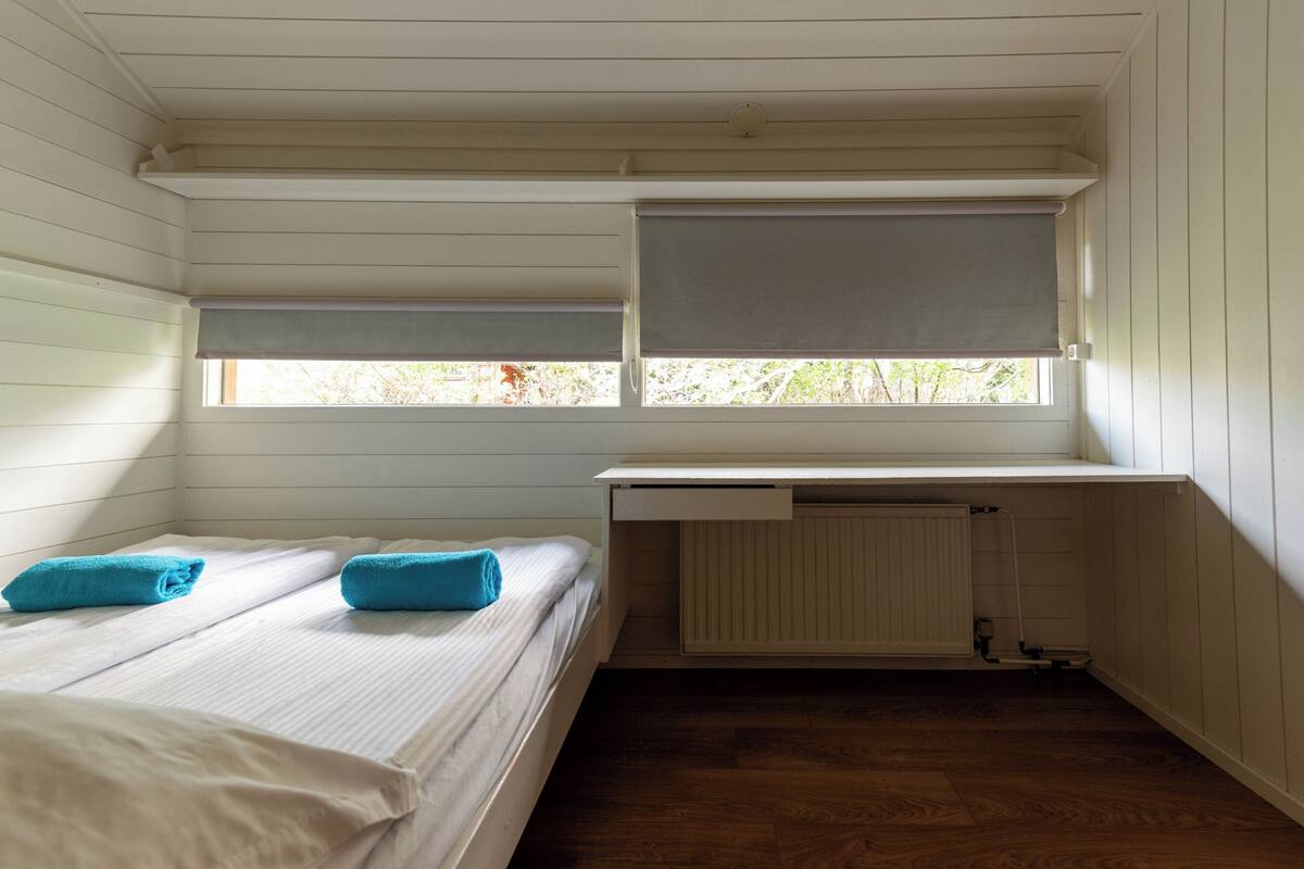 A small, single-room cabin with a bed, a window with a blind, and a radiator under a desk.