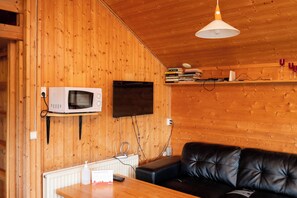 A wooden interior with a black leather sofa, a mounted TV, a microwave, and a shelf with books and glasses. - Gladheimar Cottages (Blönduós)