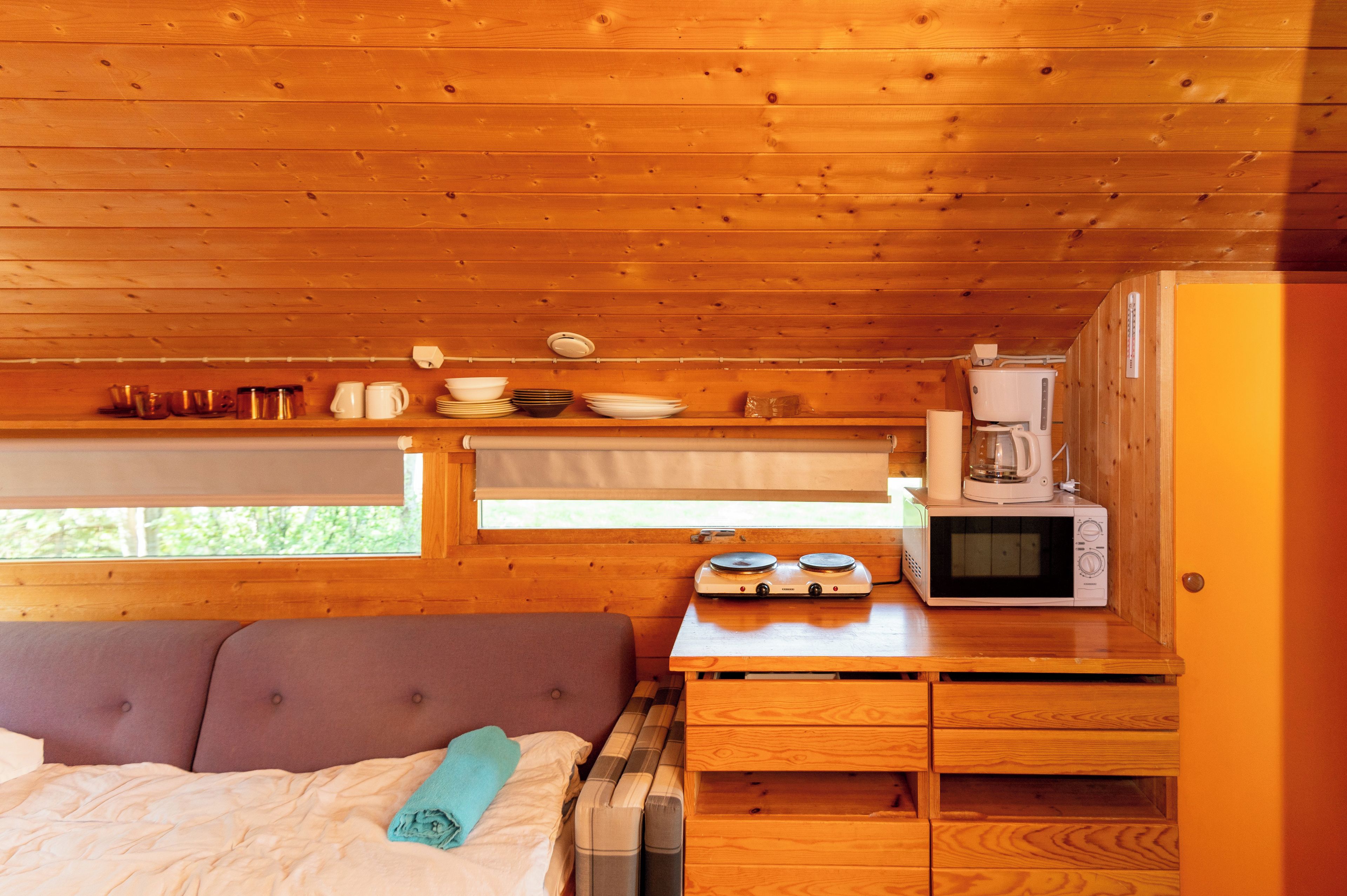 Un intérieur de cabane en bois compact, comprenant un lit, une table avec un four à micro-ondes et une machine à café, ainsi qu’une fenêtre avec des stores.