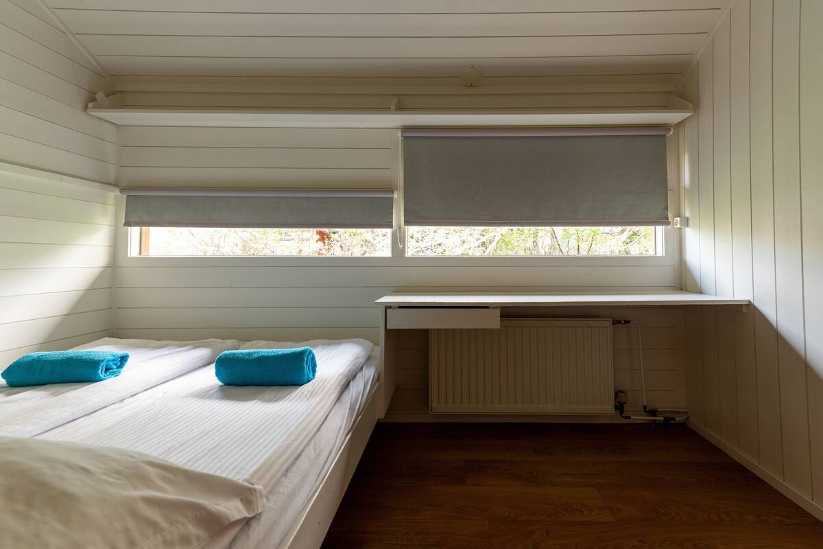 A small, single-room cabin with a bed, a window with a blind, and a radiator under a desk.