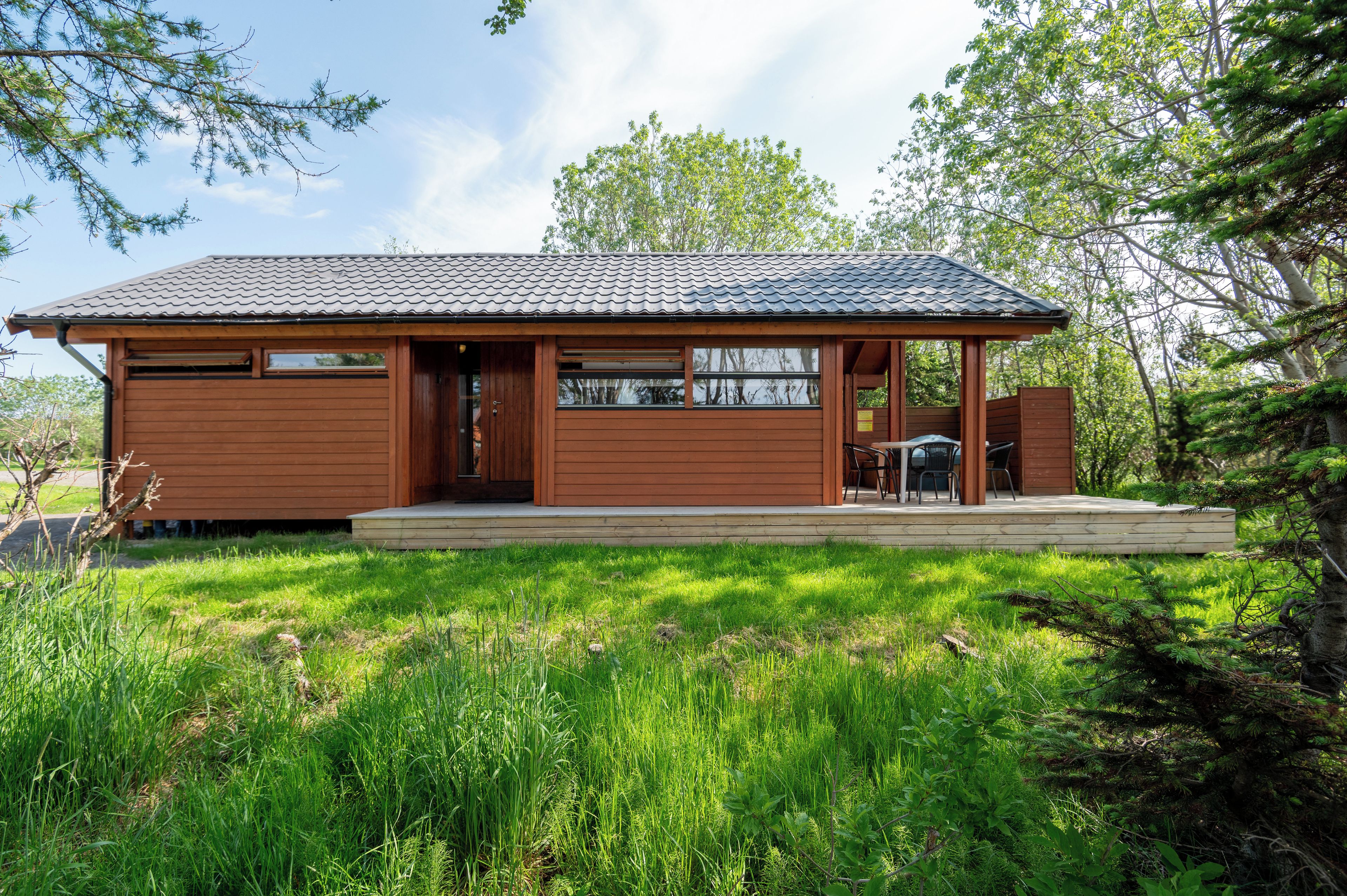 A single-story wooden cabin with a tiled roof, a front porch, and a small outdoor seating area.