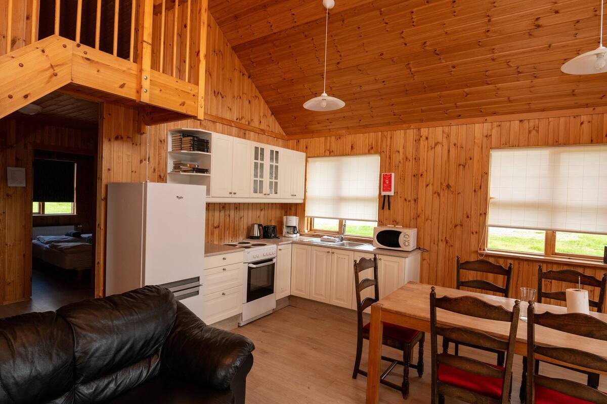 A wooden cabin interior with a kitchen, dining area, and a living room with a leather couch.