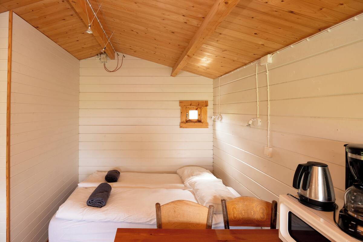 A small, cozy room with a bed, a chair, a table, and a kitchenette area with a coffee maker.