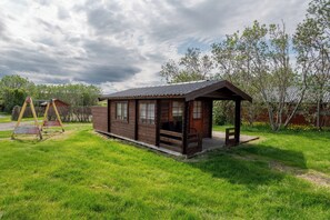 A wooden cabin with a porch, surrounded by a grassy yard and trees. - Gladheimar Cottages (Blönduós)