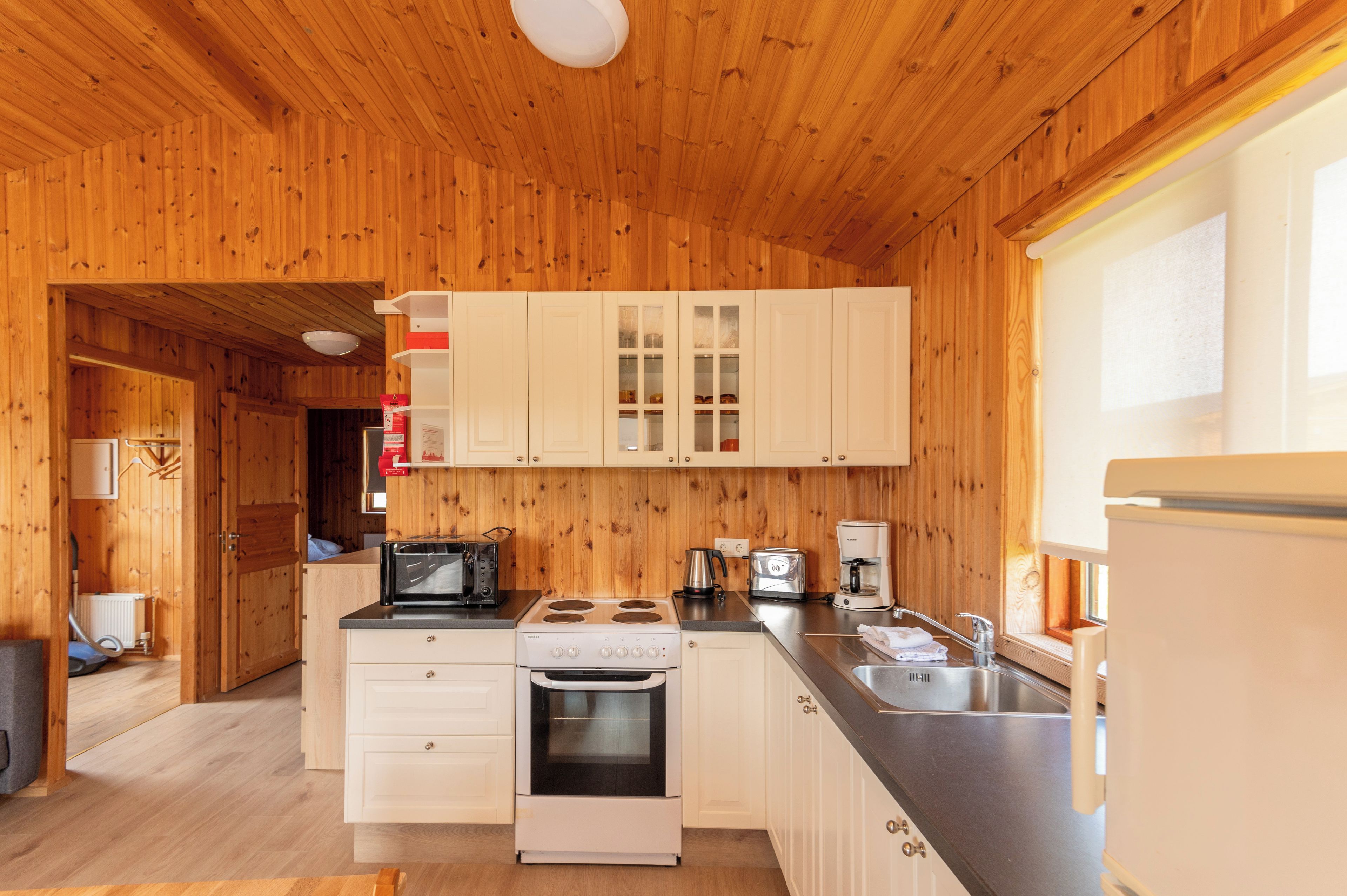 A compact kitchen with wooden walls, white cabinets, a stove, a microwave, a coffee maker, and a sink.