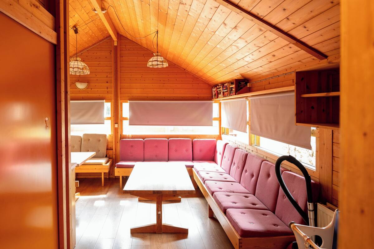 A cozy cabin interior with a wooden ceiling, a sofa, a table, and a window with a view.