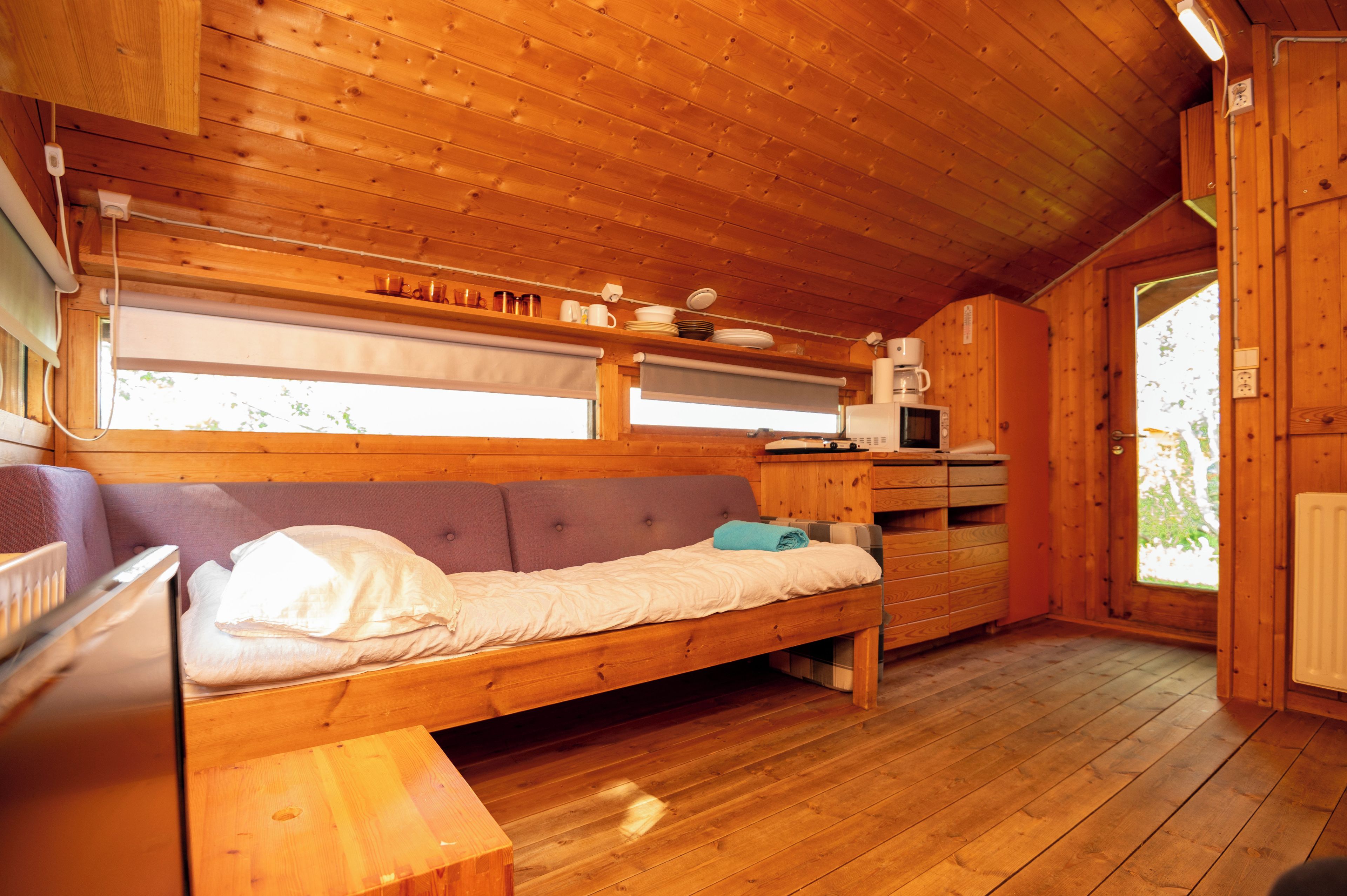 Interior de uma cabana de madeira com cama, uma mesa pequena, micro-ondas e janela com persianas.