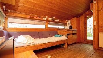 Interior de uma cabana de madeira com cama, uma mesa pequena, micro-ondas e janela com persianas.
