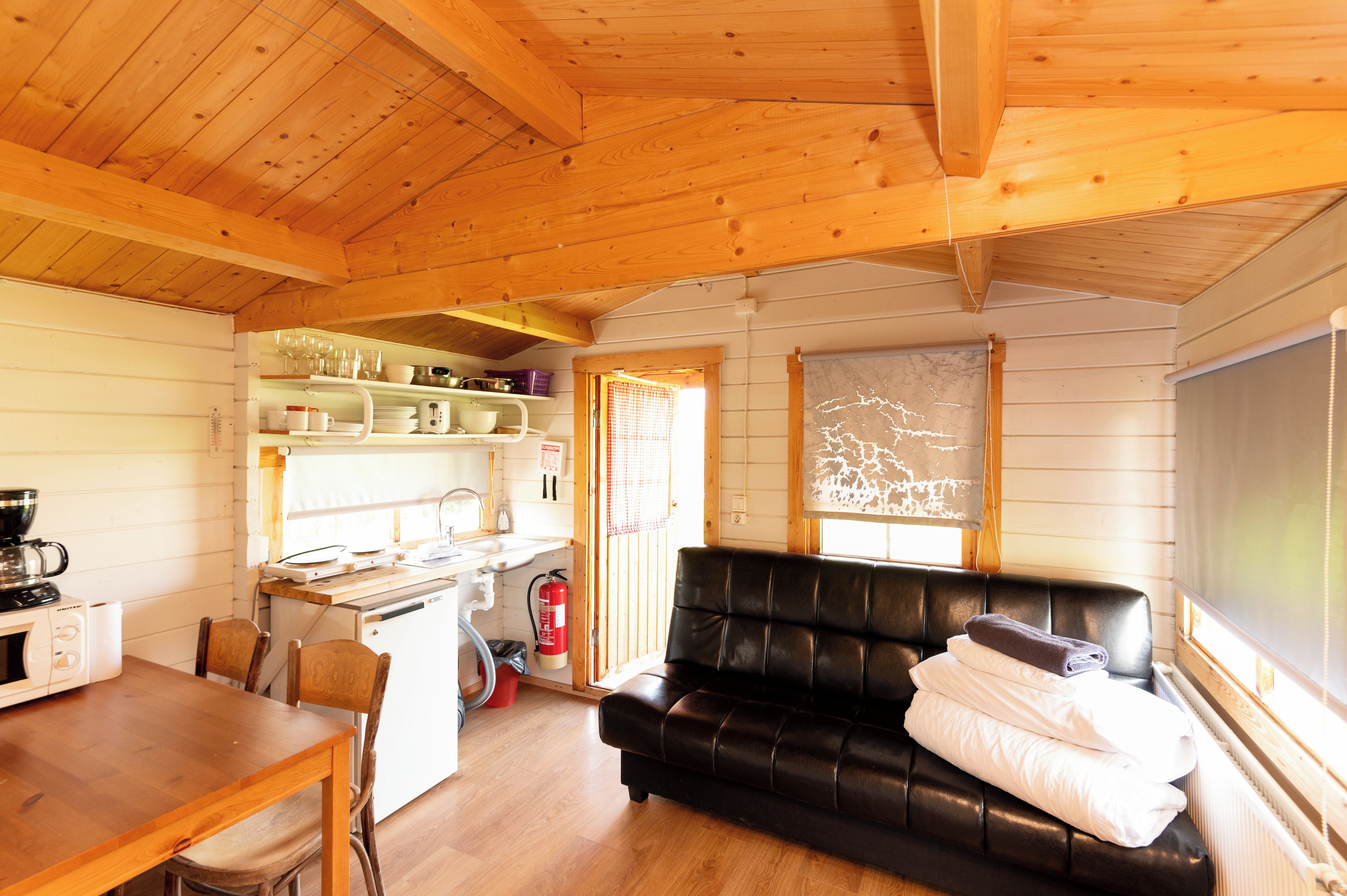 A cozy cabin interior with a kitchen area, a dining table, a black leather sofa, and a window with a view.