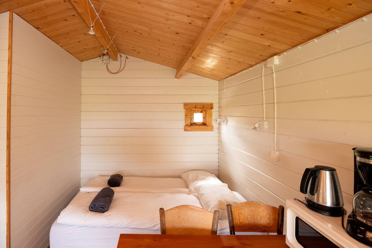 A small, cozy room with a bed, a chair, a table, and a kitchenette area with a coffee maker.