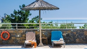 A poolside area with a thatched umbrella, two lounge chairs, a lifebuoy, and a stone wall. - Dias Luxury Studios & Apartments (Hersonissos)