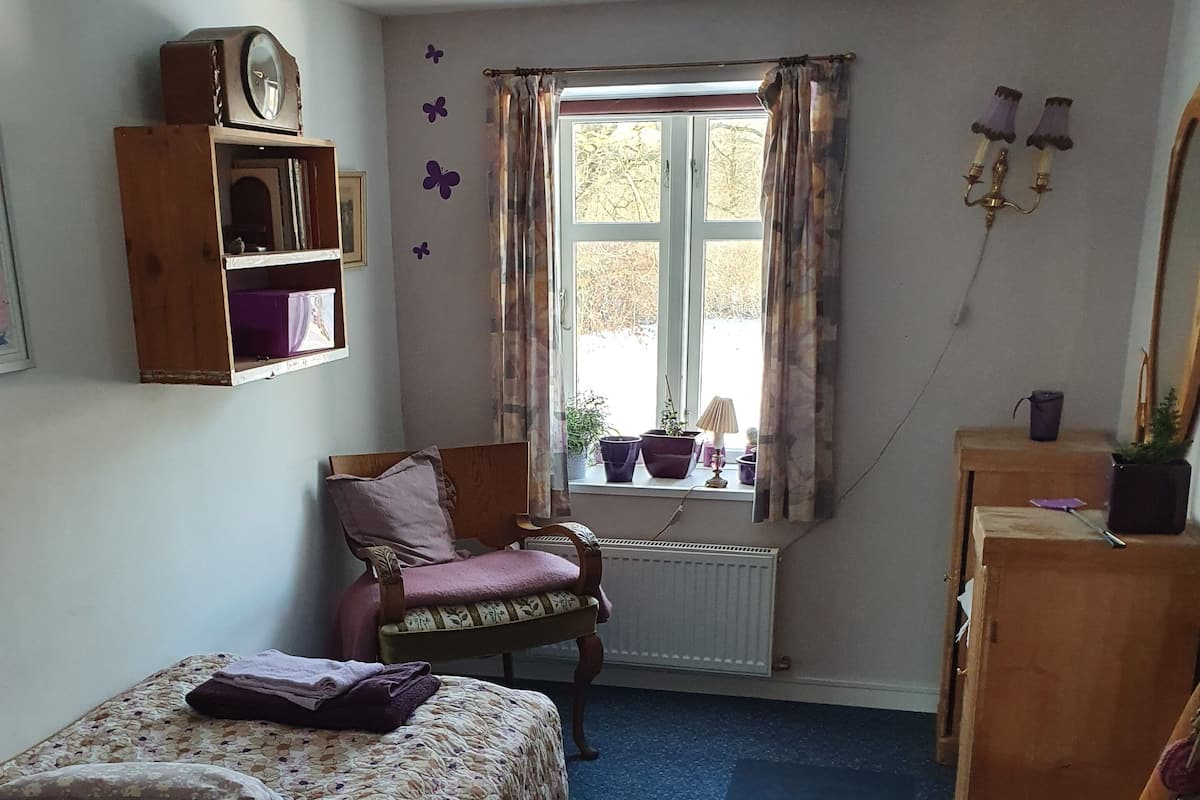 A cozy room with a bed, a chair, a wooden dresser, a window with curtains, and a radiator.