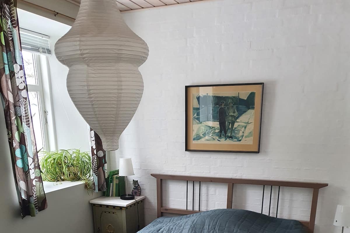 A neatly made bed with a blue quilt, two pillows, and folded towels. A framed painting hangs above the bed, and a lamp is on a bedside table.
