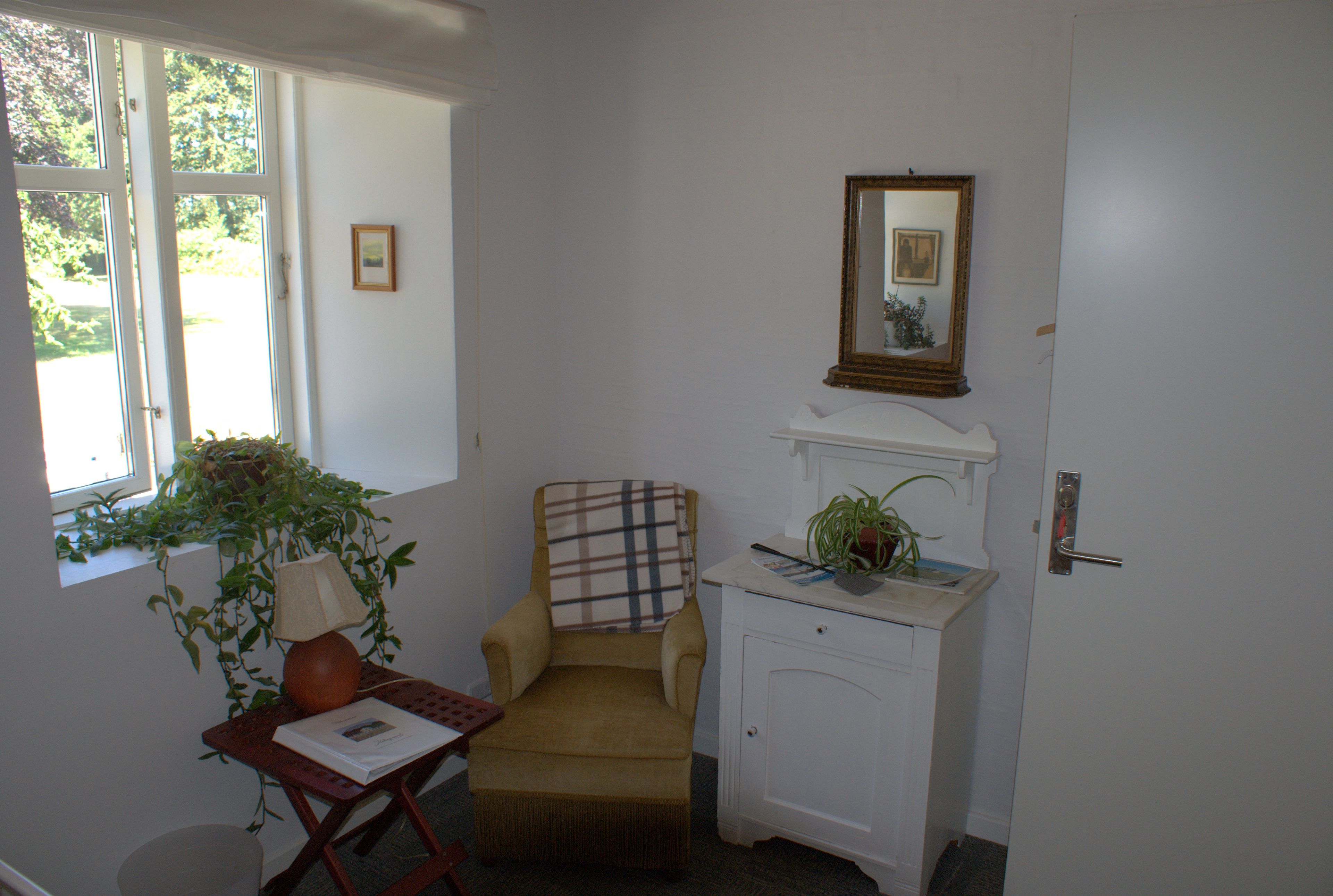 A room with a window, a chair, a desk with a lamp, a small cabinet, and a mirror on the wall.