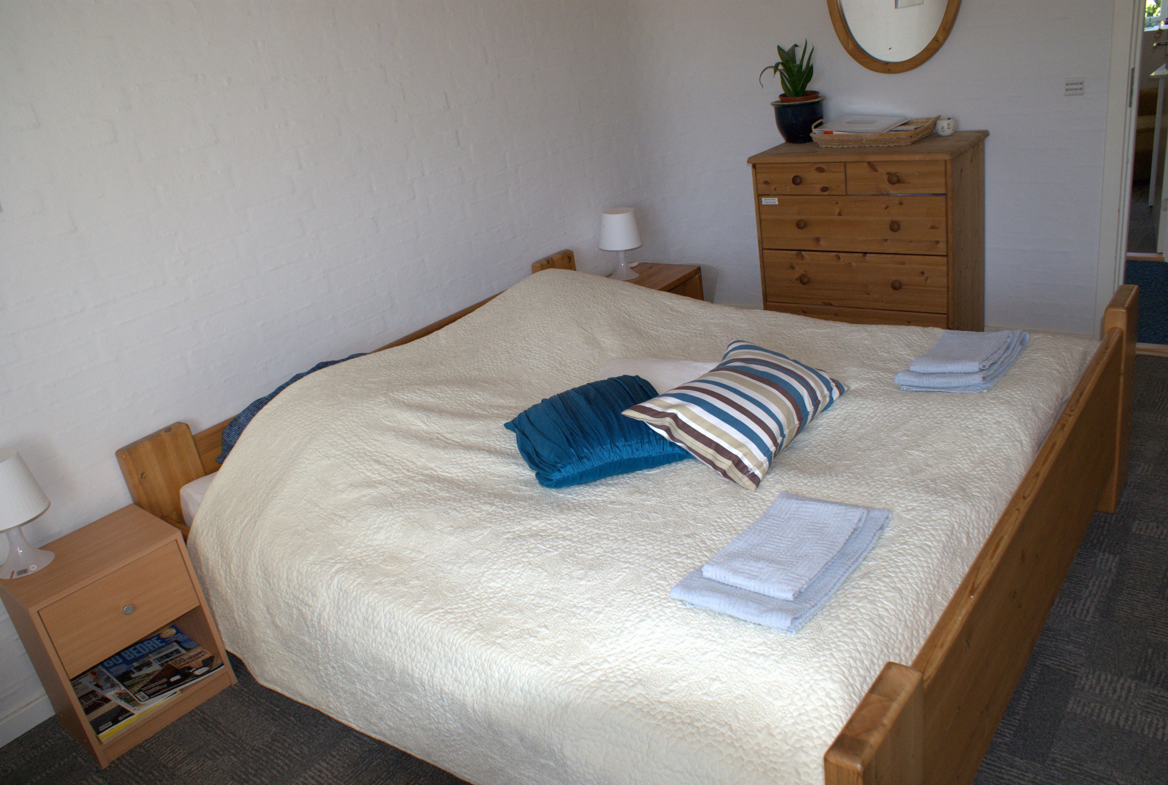 Una cama bien tendida con un cojín azul a rayas, un aparador de madera con un espejo, y una mesita de noche con una lámpara y un libro.