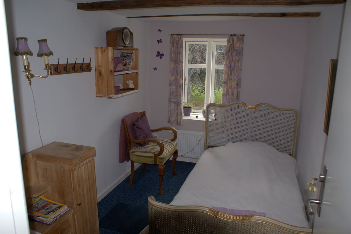 A bedroom with a single bed, a wooden chair, a small wooden cabinet, a window with curtains, and wall-mounted shelves.