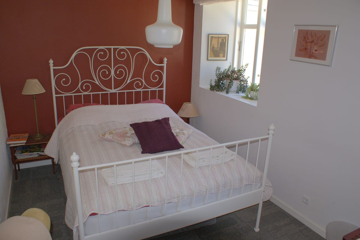 A neatly made bed with a white metal frame, a window with a view of greenery, and a bedside table with a lamp and a book.