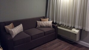 Una sala de estar con un sofá marrón oscuro, dos almohadas, un calentador portátil y una ventana con cortinas blancas.