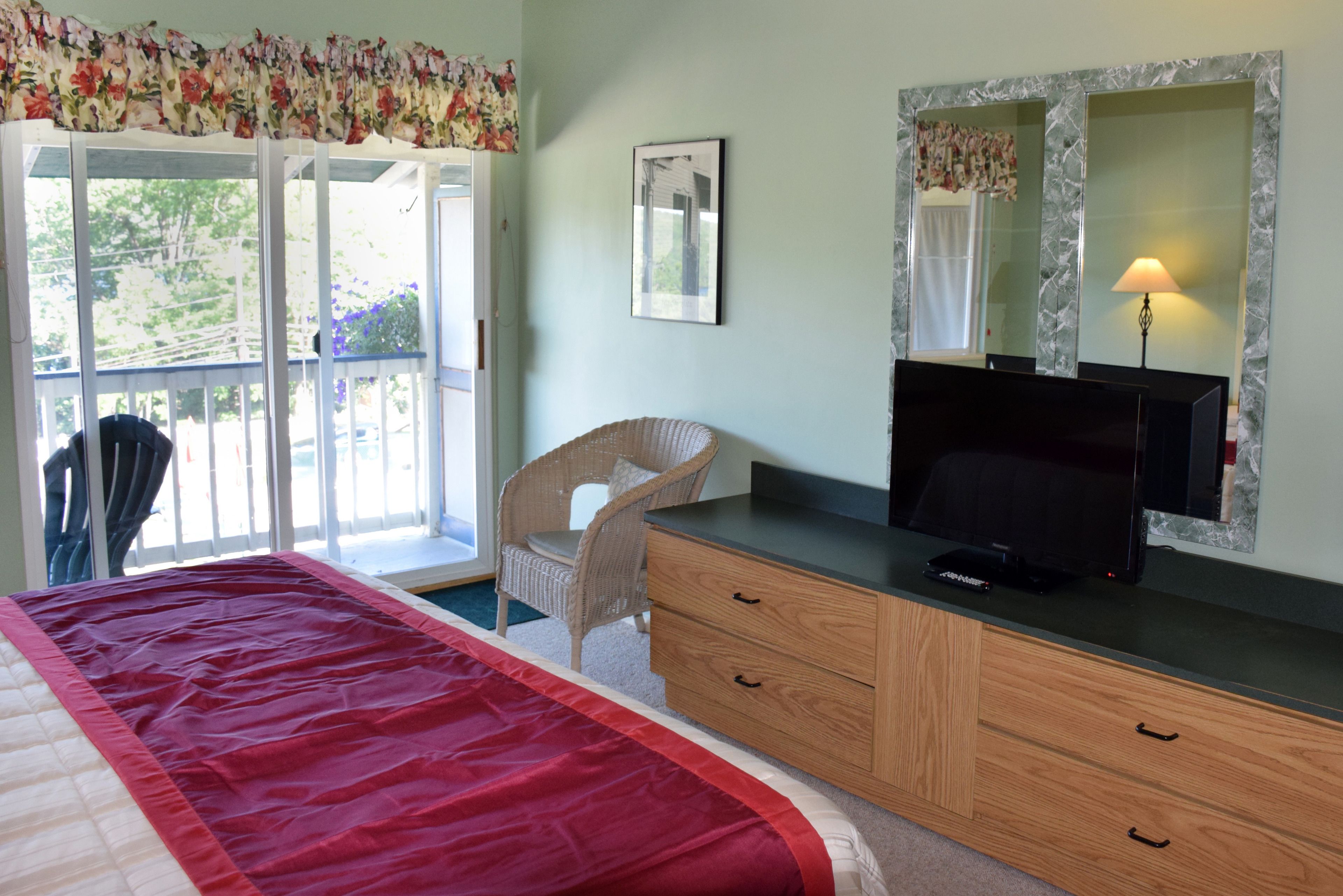 A bedroom with a bed, a dresser, a television, a chair, and a window with a view of greenery.