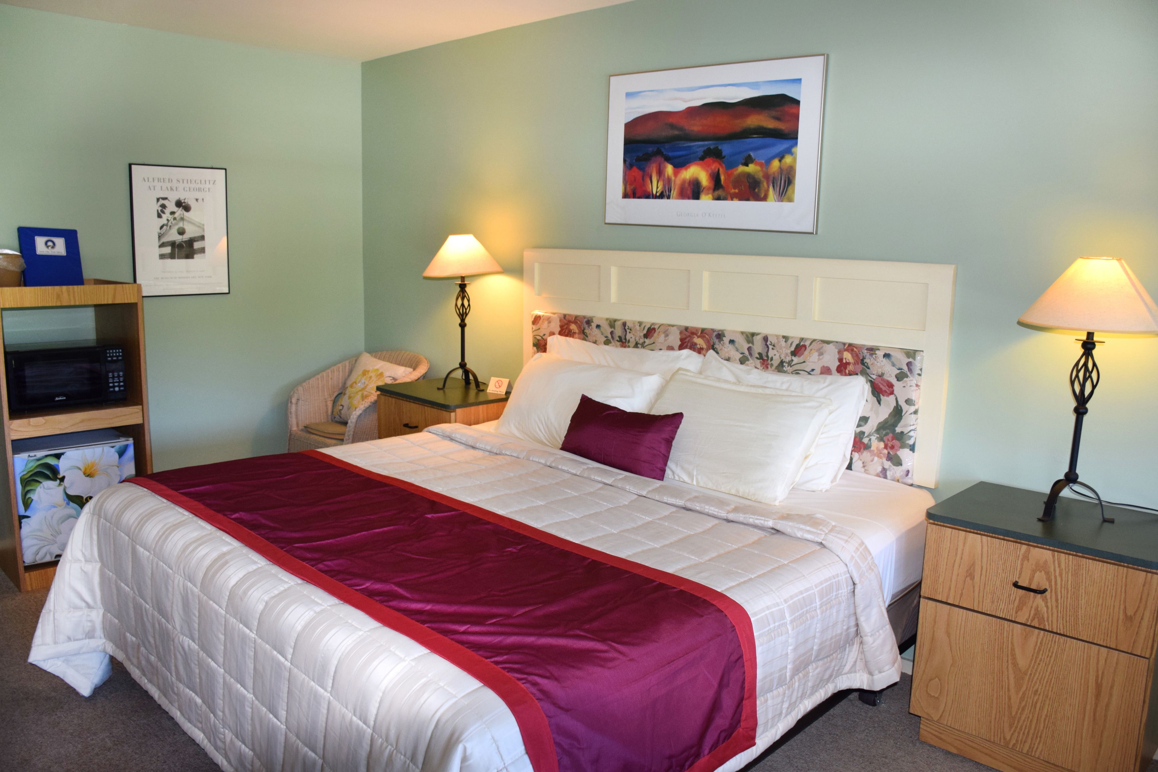 A neatly made bed with a red and white bedspread, a wooden nightstand with a lamp, and a framed picture on the wall.