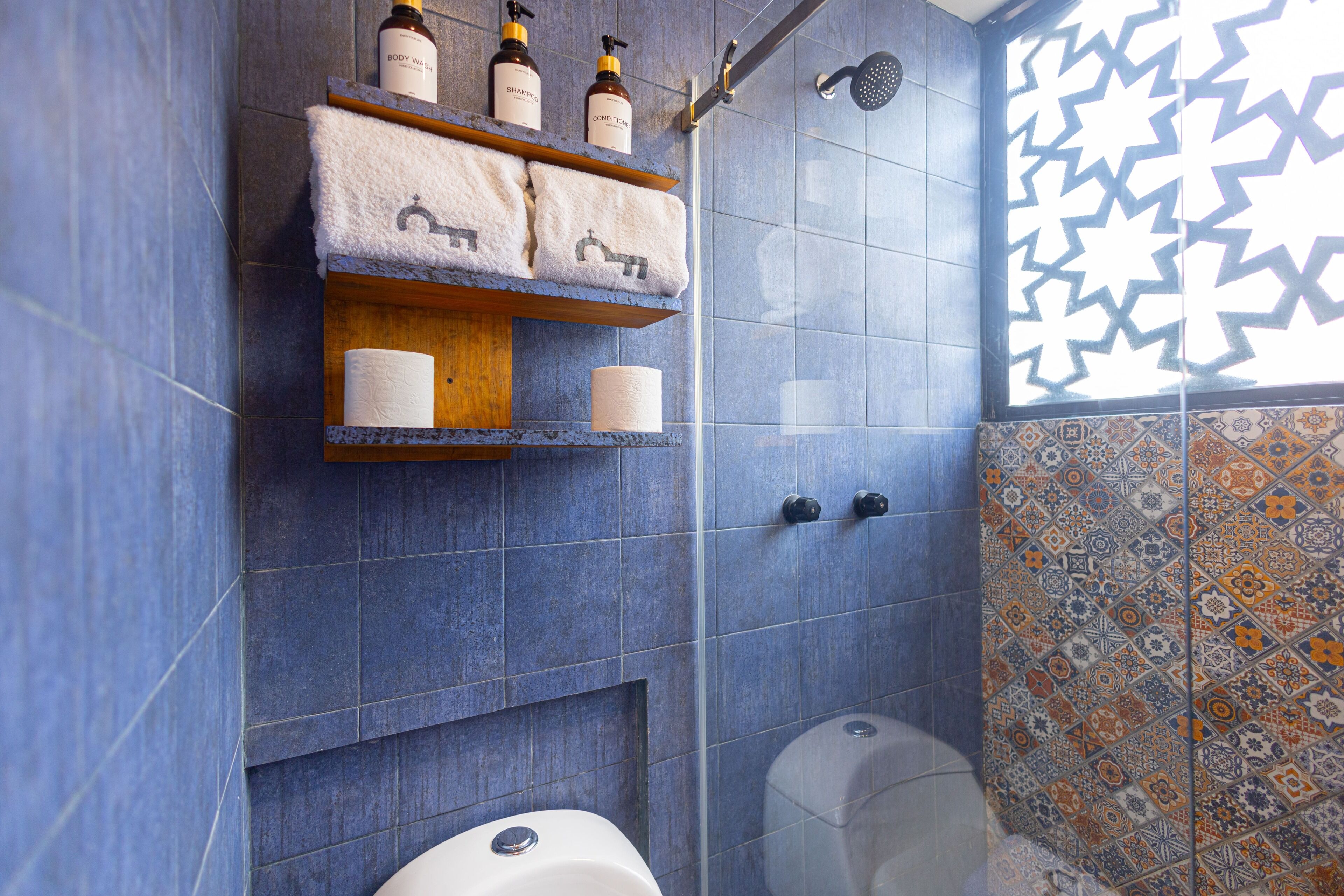 Un baño con azulejos azules, una ducha, un inodoro y un estante con artículos de aseo.