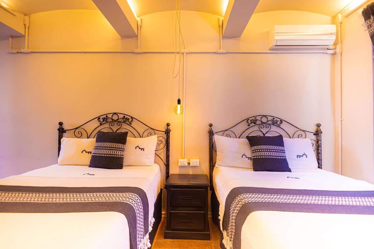 A double room with two beds, each with patterned bedding and decorative pillows. There is a small bedside table between the beds, and an air conditioning unit mounted on the wall above.
