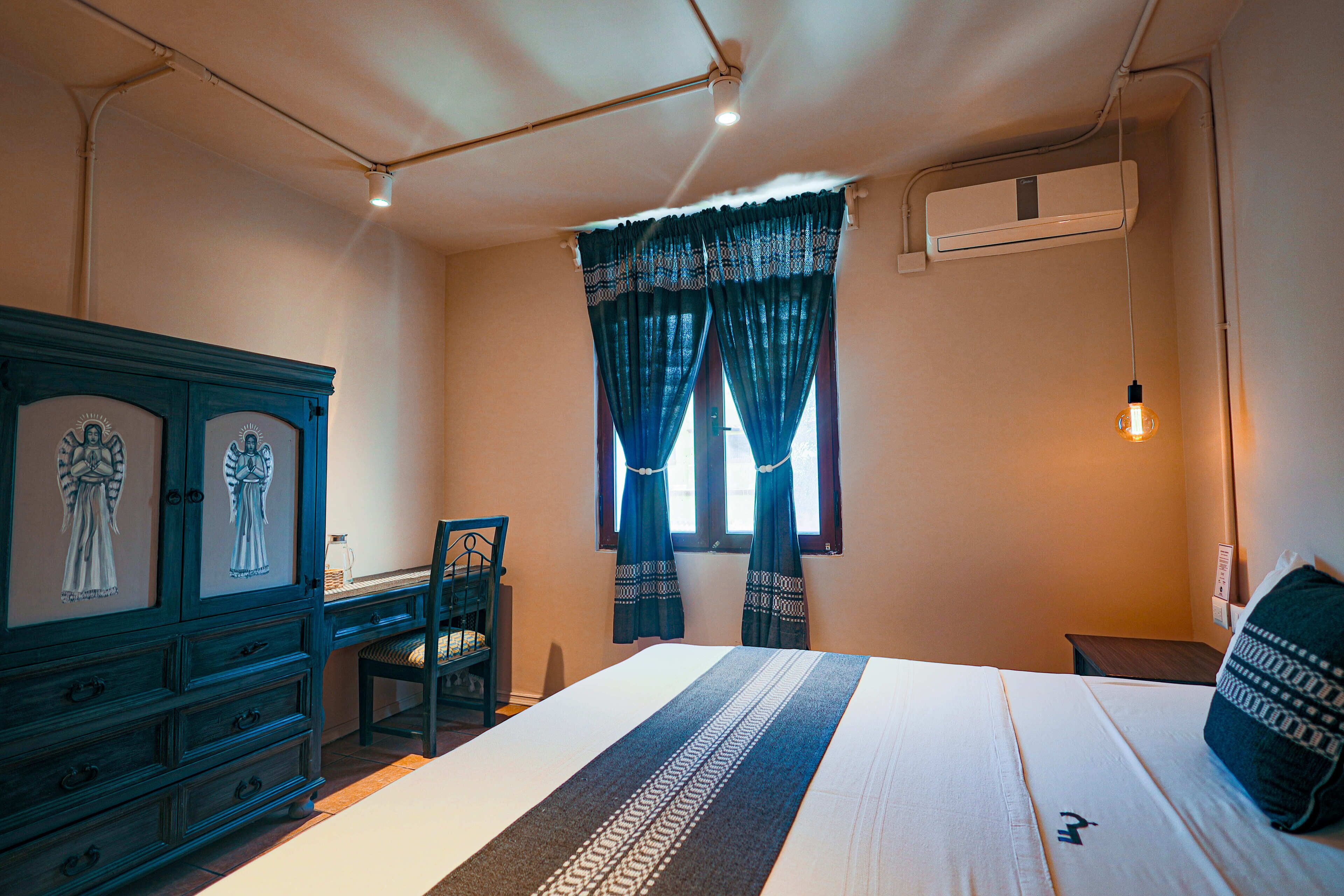 Quarto com cama, cômoda, cadeira, janela com cortinas e ar condicionado.