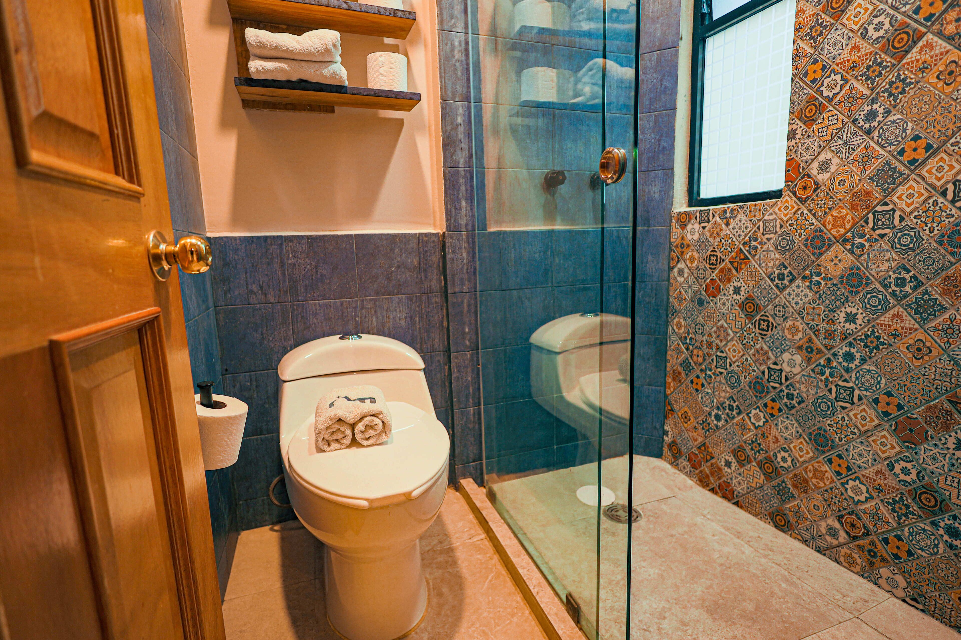 Baño con inodoro, cabina de ducha de vidrio y pared con azulejos estampados.