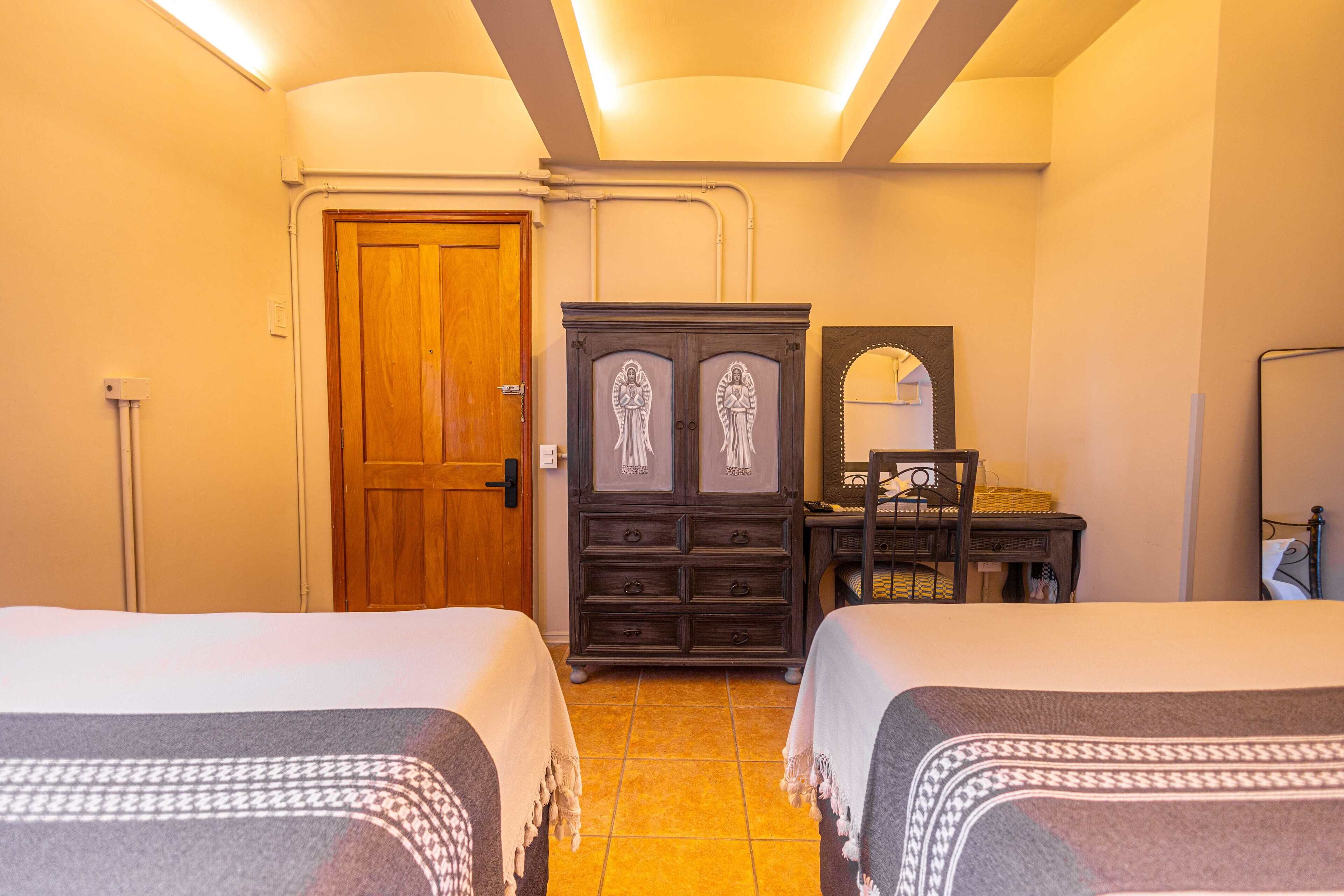 A hotel room with two beds, a wooden wardrobe, a desk with a chair, and a mirror.