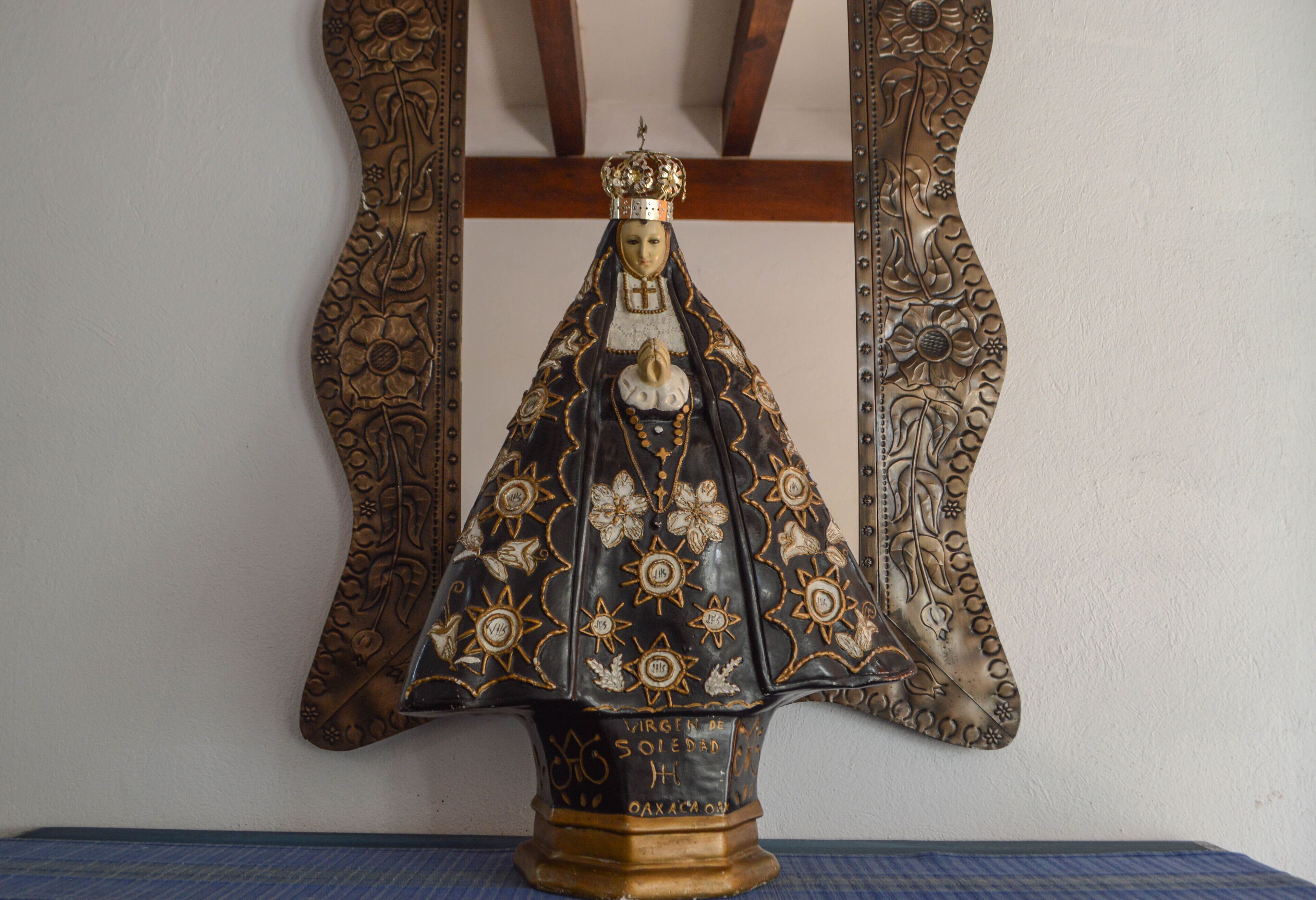 Une statue représentant une figure religieuse, ornée de motifs floraux complexes, est placée sur un piédestal portant des inscriptions, le tout adossé à un mur sobre agrémenté de poutres en bois.