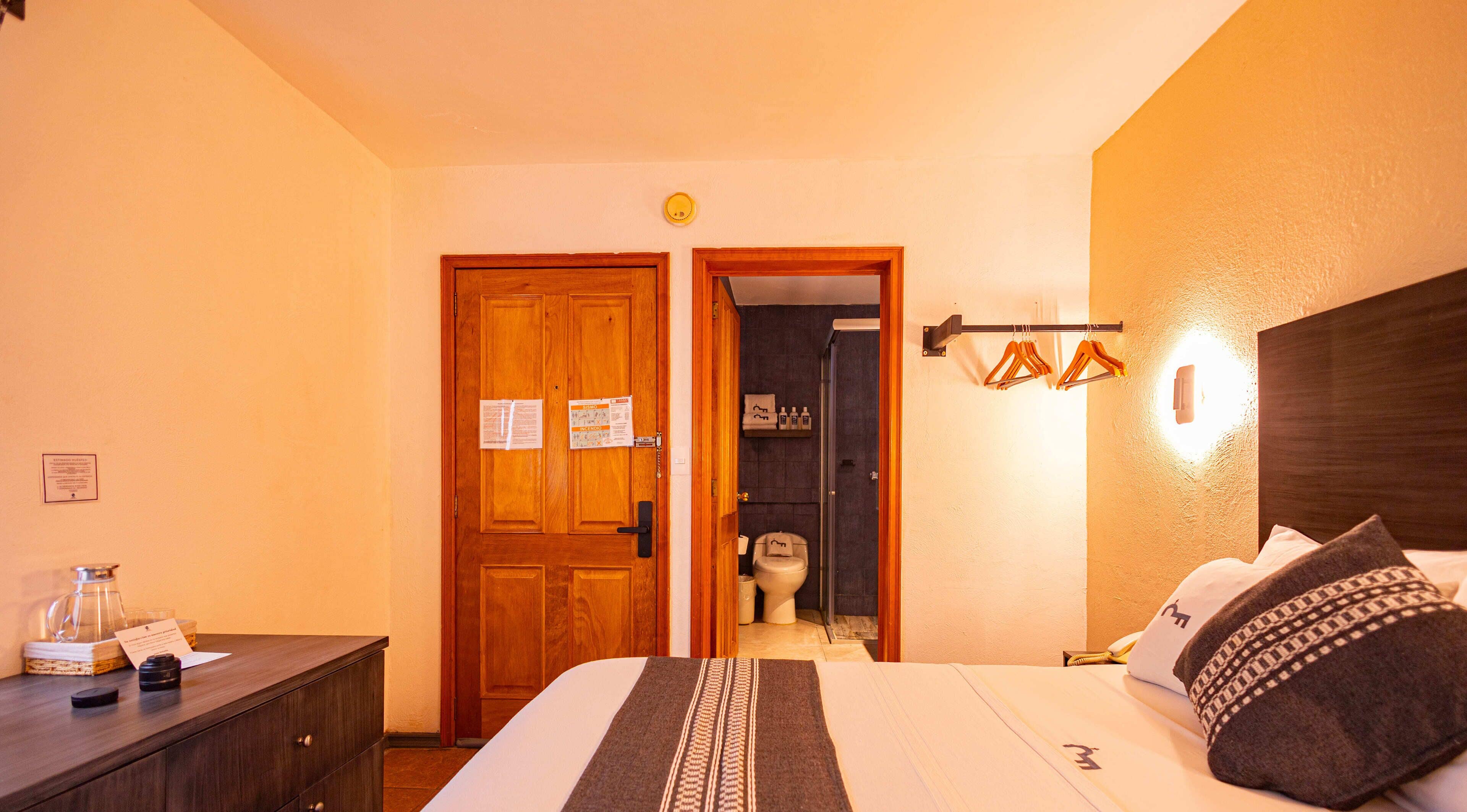 Habitación de hotel con una cama, un aparador de madera, un baño con inodoro y perchero, y una puerta con avisos.