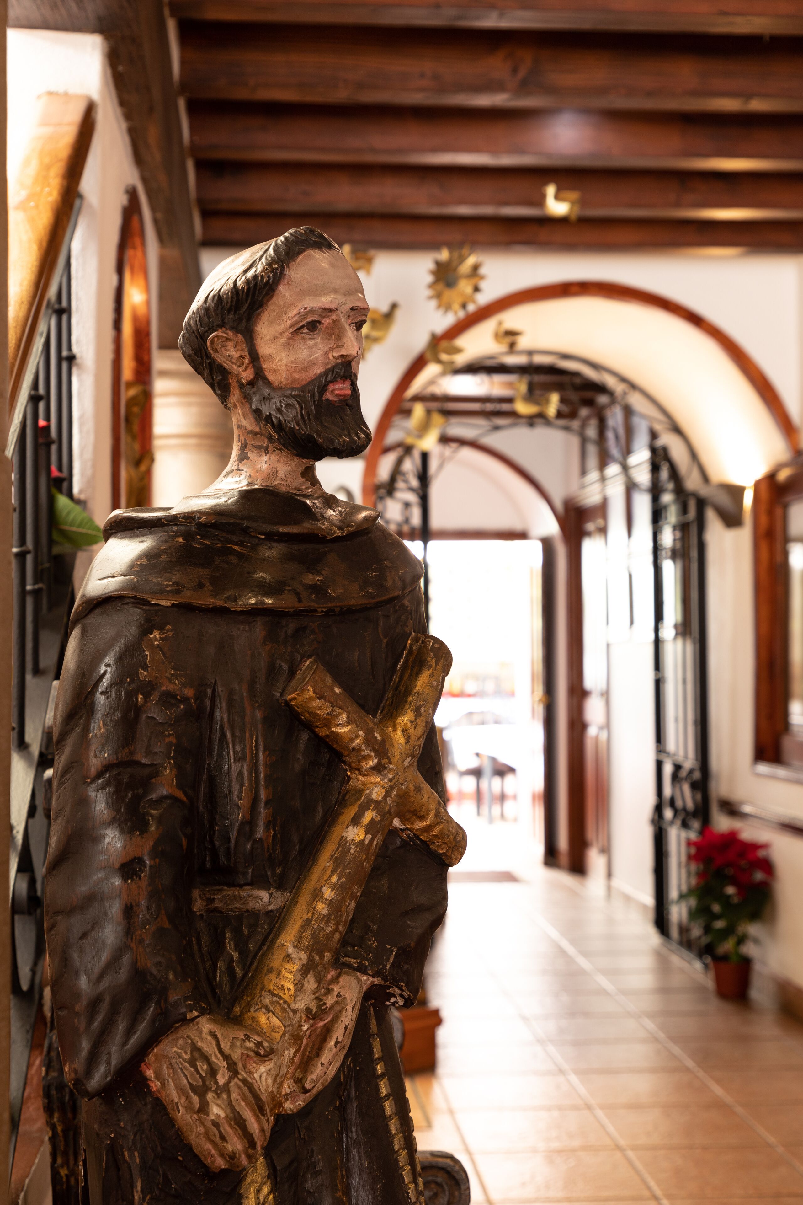Una estatua de una figura histórica sosteniendo una cruz en un pasillo con vigas de madera y un arco de entrada.