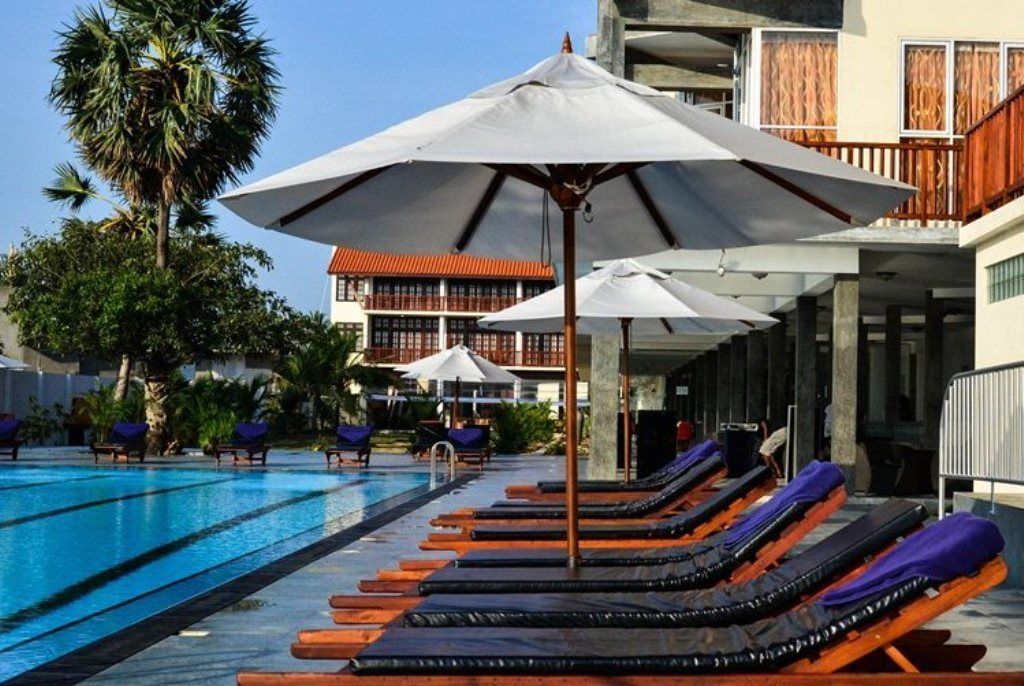 Une piscine entourée de chaises longues et de parasols, bordée de palmiers et d’un bâtiment doté de balcons.