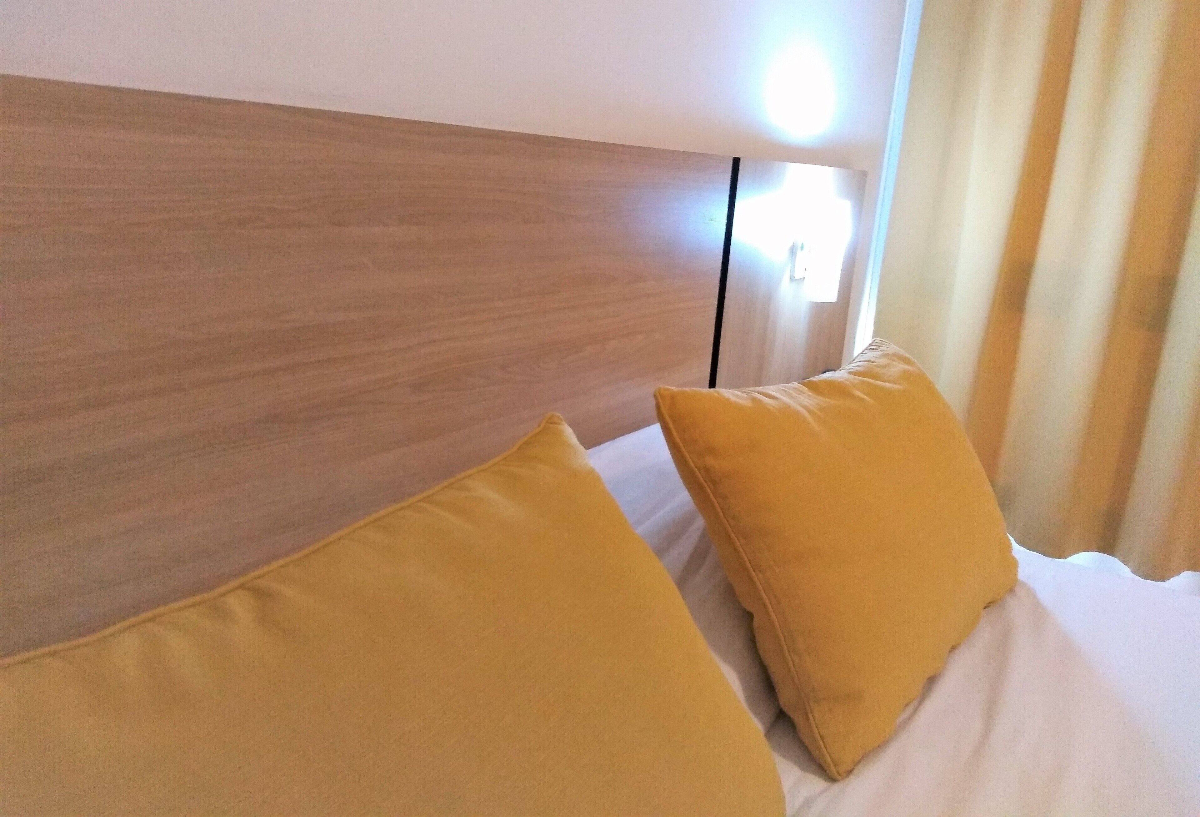 Une chambre d’hôtel avec une tête de lit en bois, deux oreillers jaunes et une couvre-lit blanche.