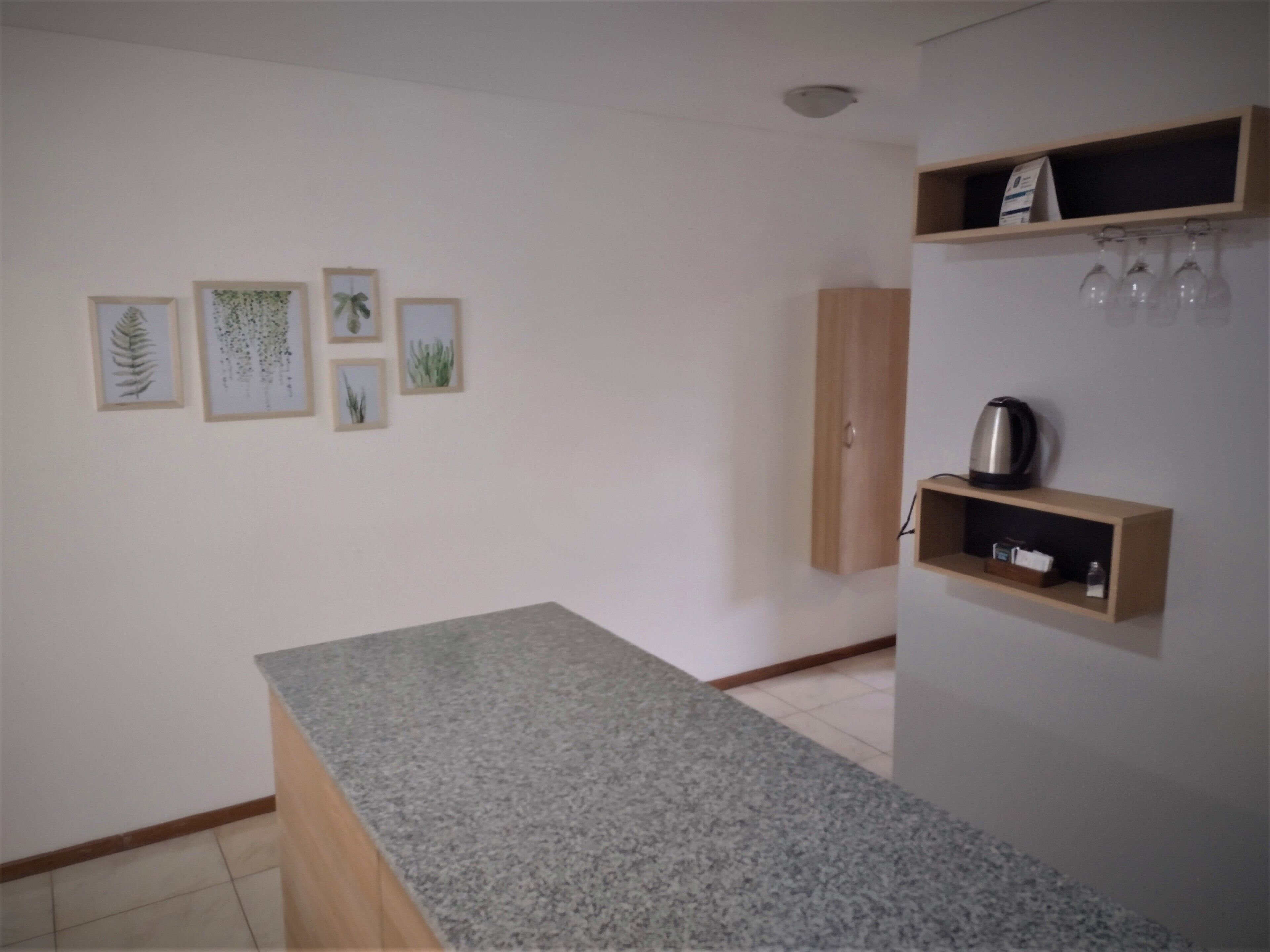 A kitchen with a granite countertop, a wooden cabinet, and a shelf with kitchenware. There are framed botanical prints on the wall.