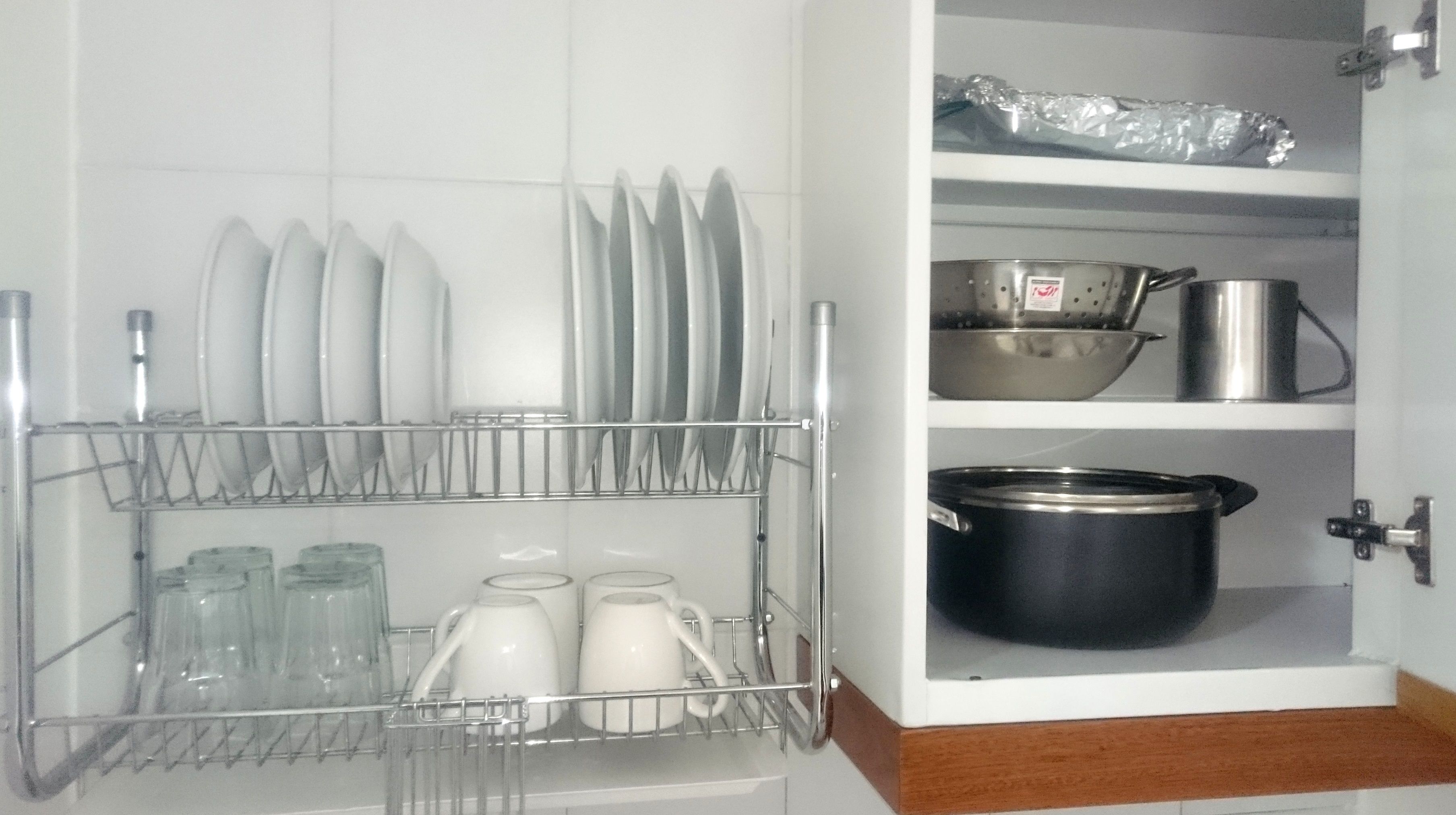 A kitchen with a dish rack holding plates and glasses, a colander, and a pot on the shelf.