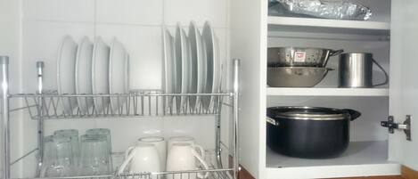 A kitchen with a dish rack holding plates and glasses, a colander, and a pot on the shelf.